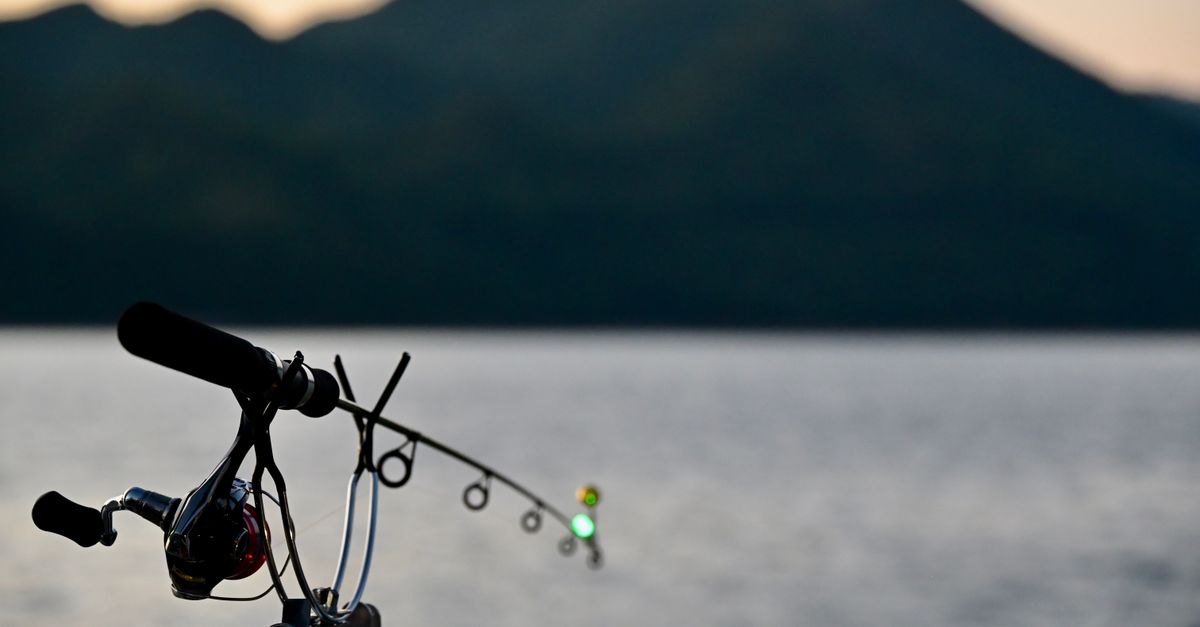 Silhouette of a fishing rod at sunset by a calm lake with mountain backdrop.